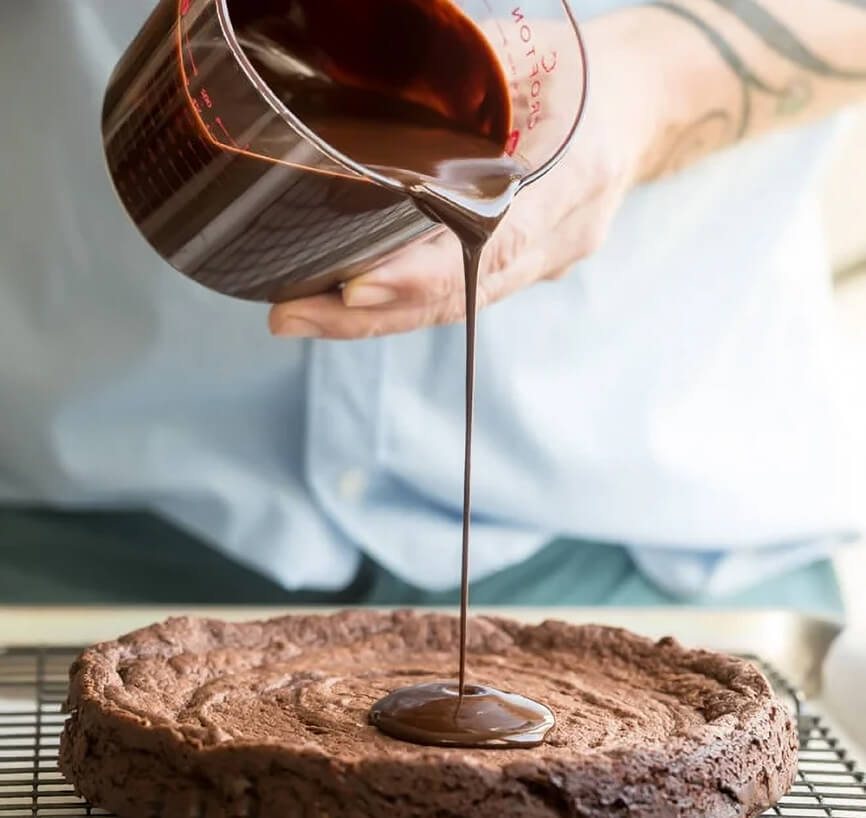 A man's hands pouring melted chocolate from a mearing cup on top of a chocolate flan that is on a cooling rack