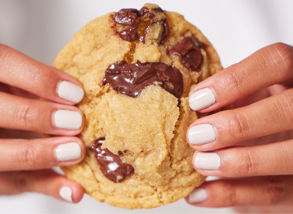 A woman's white manicured fingers hold a warm and melty chocolate chip cookie